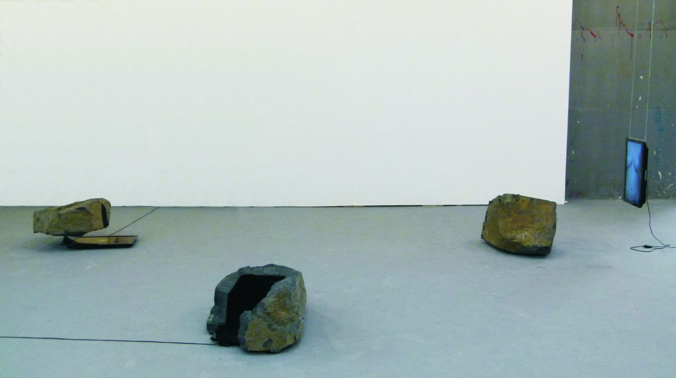 Das Stein Künstler Jan Moritz Stahl - Drei Steine auf einem grauen Boden, einer mit Kabel, daneben ein schwarzes Monitoren.