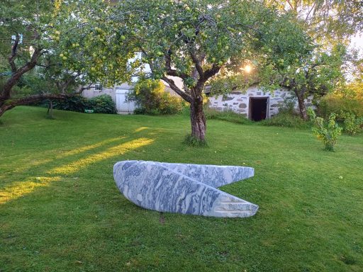 Künstler Jan Moritz Stahl - Graue Steinfigur in einem Garten mit Bäumen und einem alten Gebäude im Hintergrund.