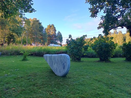 Künstler Jan Moritz Stahl - Grauer, geschwungener Stein auf einer Wiese, umgeben von Bäumen und Sträuchern.