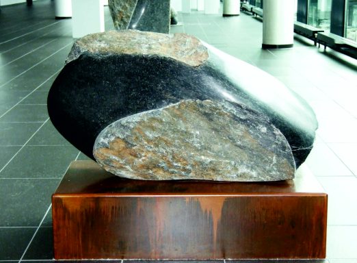 Künstler Jan Moritz Stahl -Abgerundeter Stein mit farbigen Schichten, auf einem Holzsockel platziert.