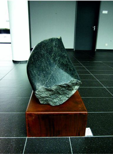 Künstler Jan Moritz Stahl - Dunkler Steinblock auf einem Holzsockel in einem modernen Innenraum.