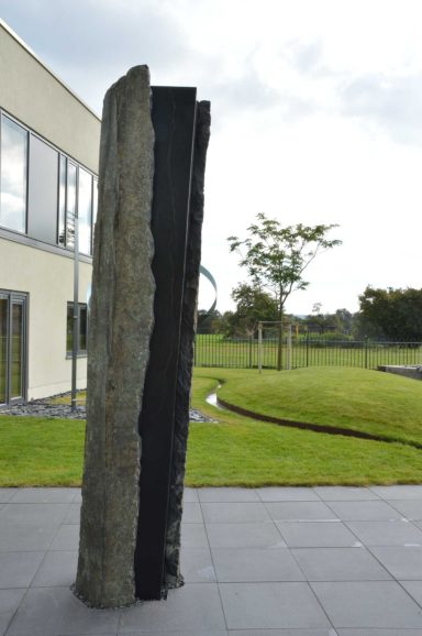 Künstler Jan Moritz Stahl - Ein hoher, schmaler Stein mit rauer Oberfläche steht im Freien vor einem modernen Gebäude.